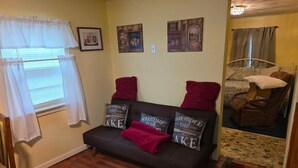 Ein gemütliches Wohnzimmer mit einer dunkelgrauen Couch, roten Kissen und einer roten Decke. An der Wand hängen zwei gerahmte Bilder, ein Fenster mit weißen Vorhängen und ein gemustertes Teppich.