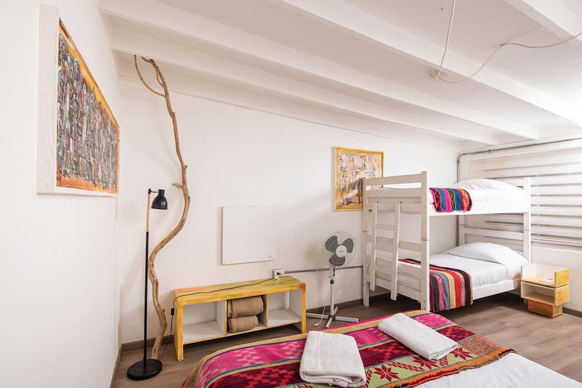 Un dormitorio con literas, un escritorio, una silla, un ventilador de pie y obras de arte en las paredes.