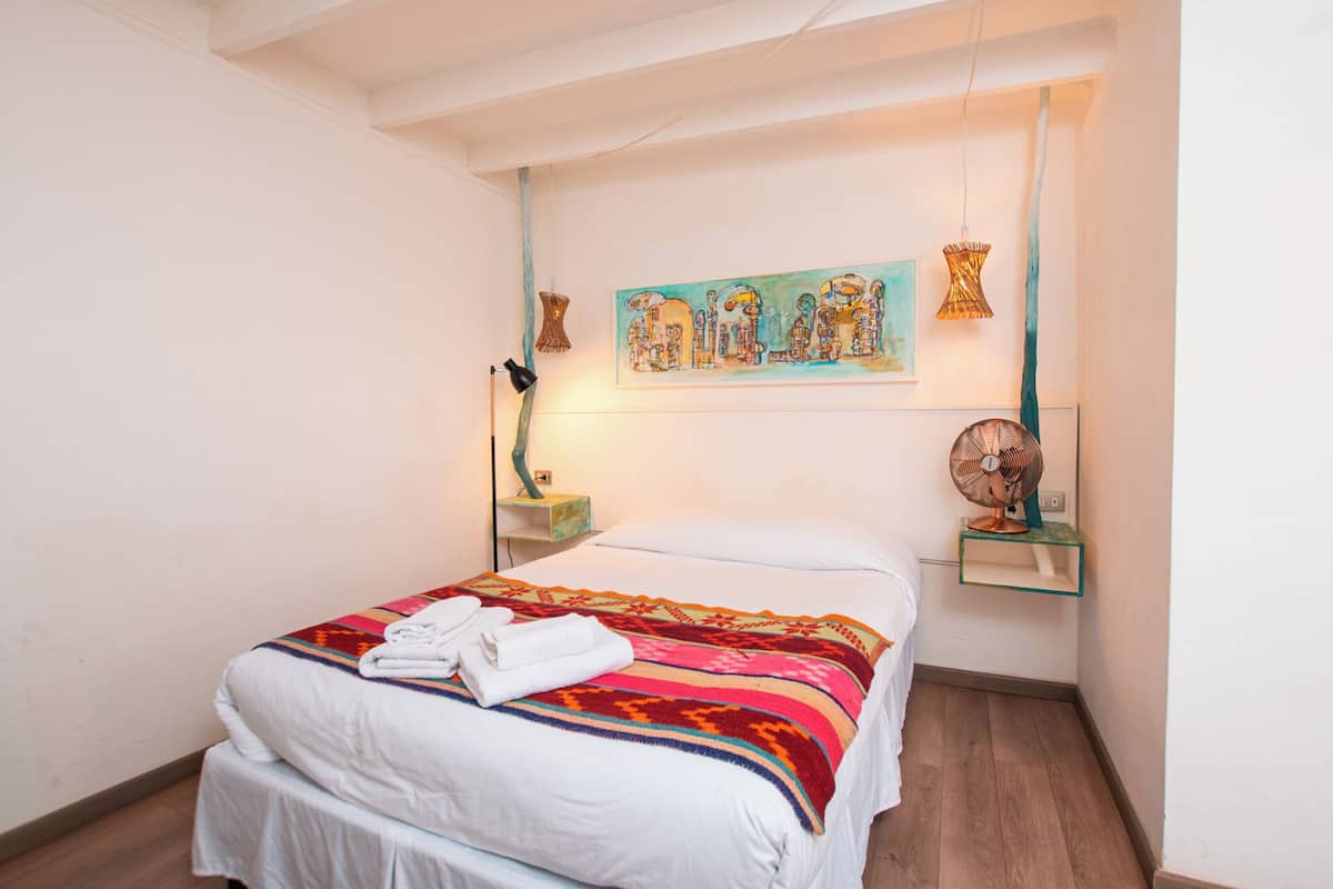 Una cama bien hecha con un edredón colorido y una toalla doblada, un cuadro colgado en la pared, una lámpara de pie y una mesita con un ventilador.