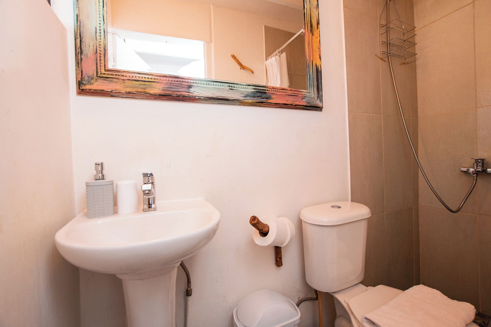 Une salle de bain avec un lavabo, des toilettes et une douche, le tout de couleur blanche. Un miroir avec un cadre coloré est placé au-dessus du lavabo.