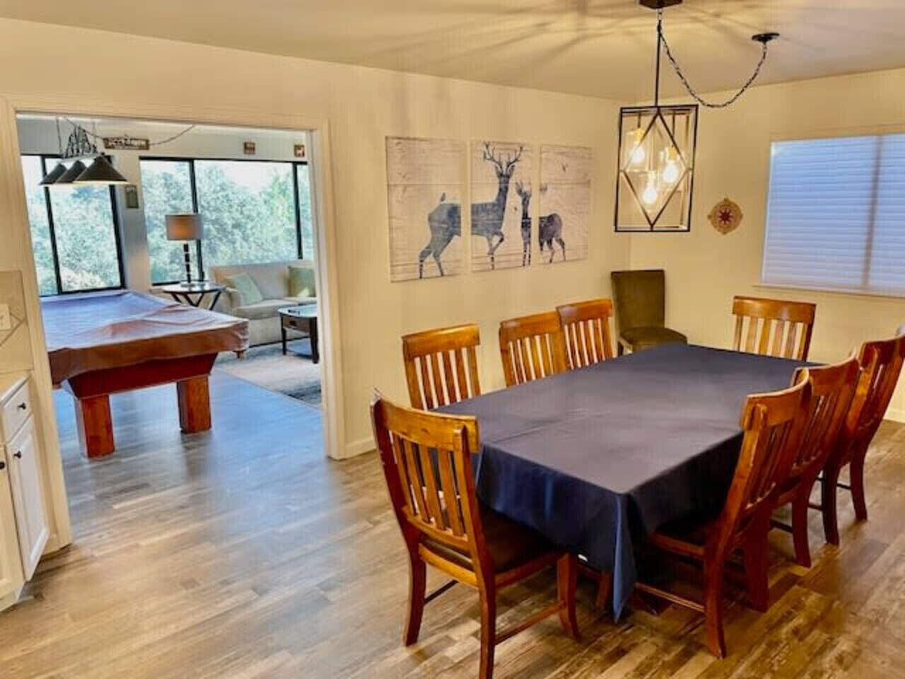 A dining area with a rectangular table, wooden chairs, and a bench. There are wall art pieces depicting deer, a chandelier, and a window with blinds.