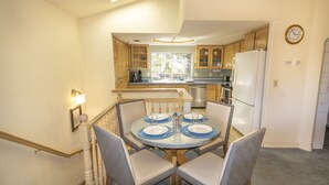 A dining area with a round table set for four, surrounded by chairs. A staircase with a railing leads to an upper level. The kitchen area is visible in the background with wooden cabinets and a refrigerator. - Squirrel View - Hot tub/BBQ/Patio/Shuffle Board (Oakhurst)