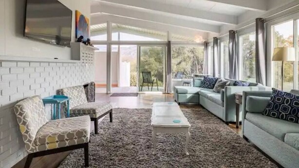A modern living room with a fireplace, a flat-screen TV mounted on the wall, and a large window providing a view of the outdoors. - Black Bear Suite/Hot Tub/Sleeps 4/affordable! (Oakhurst)