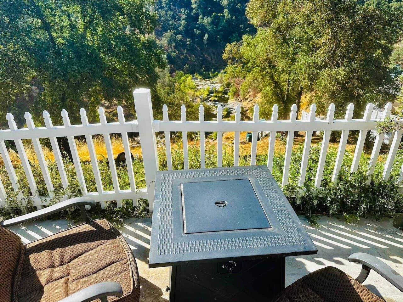 A patio with a table and chairs, a white picket fence, and a view of a river and forest.