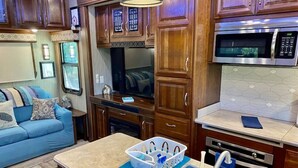 A compact RV kitchen with wooden cabinets, a microwave, a sink, and a dining area with a blue couch. - Happy Hedgehog - River Views - Hot Tub - Private (Ahwahnee)