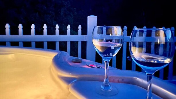 A hot tub with two glasses of white wine, surrounded by a white fence and a mountainous backdrop. - Happy Hedgehog - River Views - Hot Tub - Private (Ahwahnee)