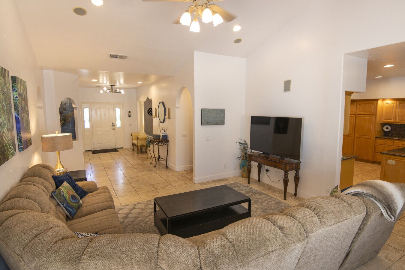 A spacious living room with a large sofa, a coffee table, and a flat-screen TV. There is a dining area with a table and chairs, and a kitchen with wooden cabinets.