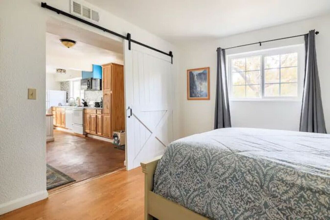 A bedroom with a bed, a window with curtains, and a view of a kitchen and living area.