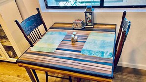 Una mesa de comedor de madera con superficie de vidrio, rodeada de sillas, en una habitación con piso de madera y una ventana al fondo.