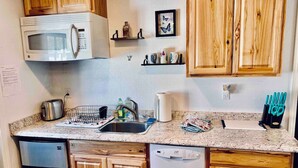 Cocina compacta con armarios de madera, refrigerador de acero inoxidable, microondas, lavavajillas y un lavamanos. Hay repisas con objetos decorativos y un bloque para cuchillos en la encimera.