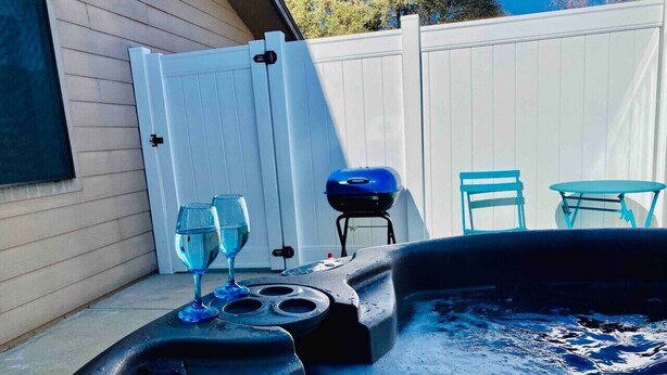 Tina de hidromasaje con un vaso y una parrilla barbacoa en un patio trasero con cerca blanca y mobiliario exterior.
