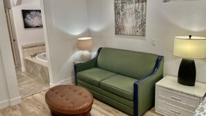 Una sala moderna con un sofá verde, un reposapiés marrón, una mesita de noche blanca con una lámpara, y un baño a la vista al fondo.