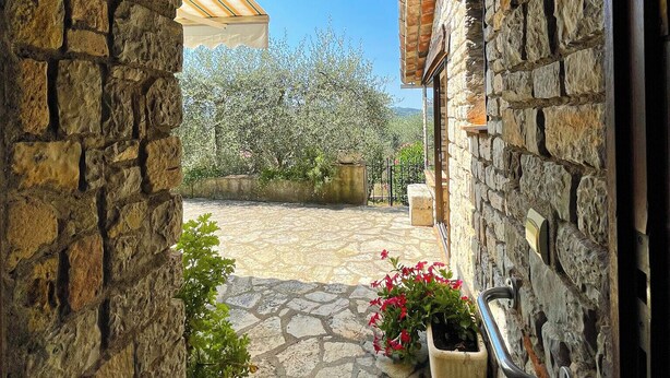 Un patio con paredes de piedra, una pérgola de madera, plantas en macetas y vistas a una zona arbolada.