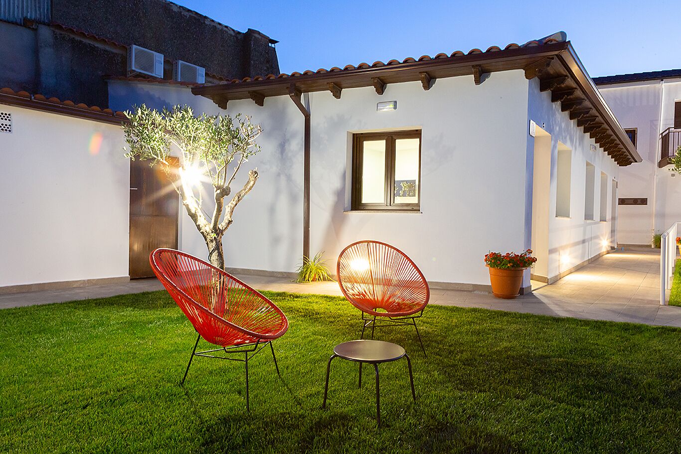 Una casa moderna con fachada blanca, techo de tejas y un pequeño jardín con dos sillas rojas y un árbol.