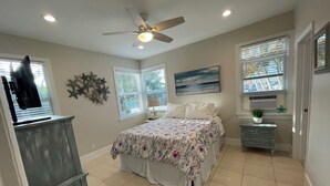 Une chambre à coucher avec un lit, un ventilateur de plafond, un bureau avec un ordinateur et une fenêtre avec des stores.