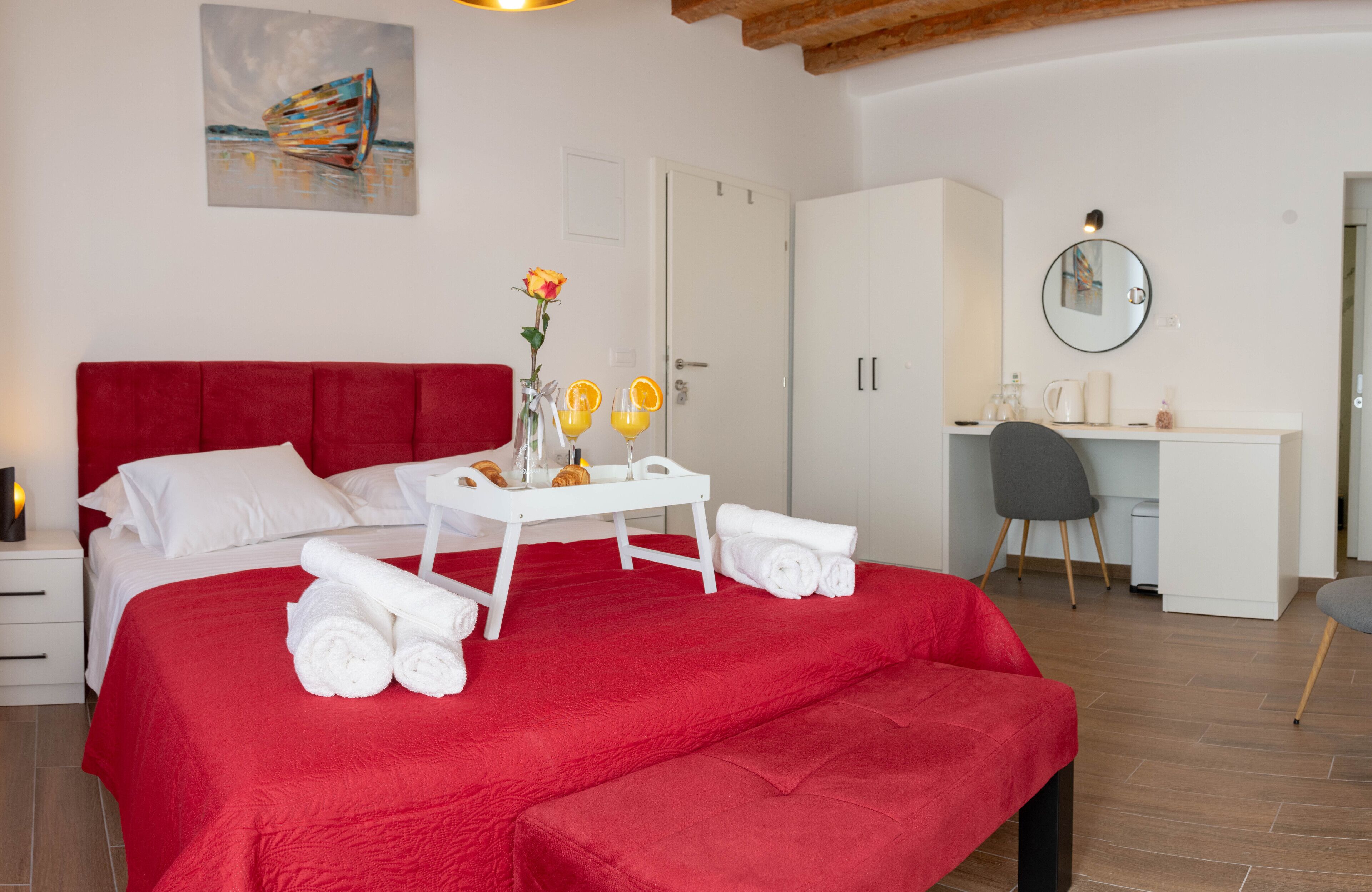 A hotel room with a red bedspread, white towels, a small table with orange drinks, a vase with flowers, and a desk with a chair.