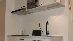 Cocina compacta con microondas, tostadora y hervidor sobre la encimera, un lavabo con grifo, y una pequeña estantería con una planta y un rollo de papel de cocina.
