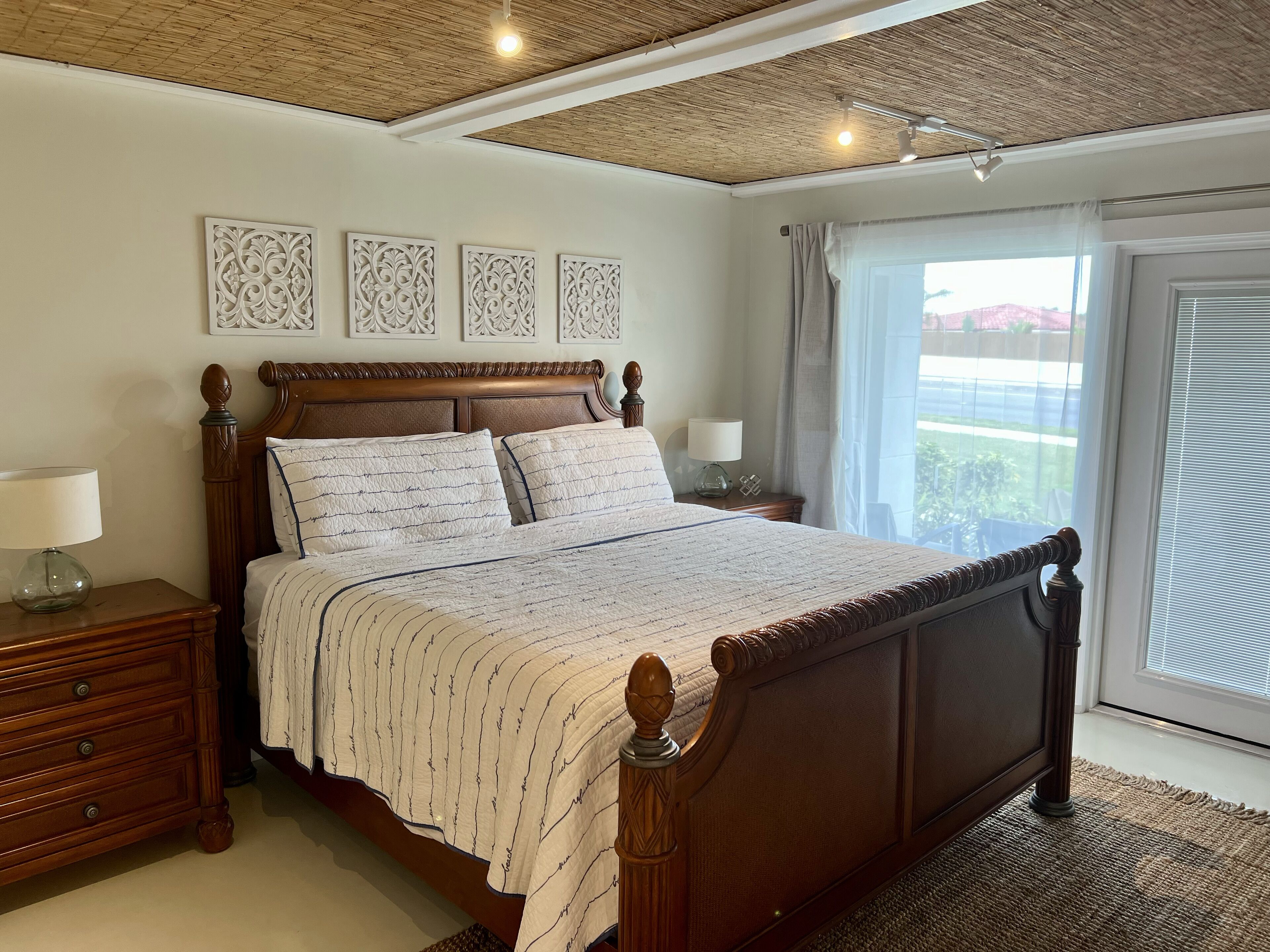 Un dormitorio con una cama de madera, paneles decorativos en la pared, una mesita de noche con lámpara y vistas al exterior a través de una ventana.