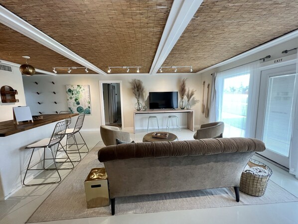 Uma sala de estar moderna com sofá, cadeiras, mesa de centro e televisão. Teto de madeira e uma janela grande.