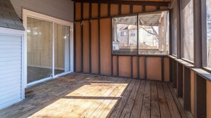 A covered patio area with wooden flooring, a sliding glass door, and a window overlooking a residential area.