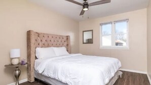 A bedroom with a tufted headboard, a bed with white bedding, a round table with a lamp, a ceiling fan, and a window with blinds.