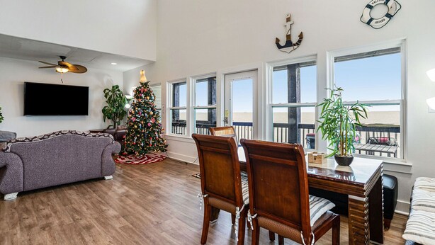Uma sala de estar espaçosa com uma televisão de tela plana grande, uma árvore de Natal, mesa de jantar de madeira com cadeiras e grandes janelas com vista para a praia.