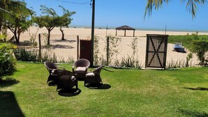 Un espace verdoyant avec des chaises en osier et une structure en bois, une zone de plage clôturée et une vue dégagée sur la plage.