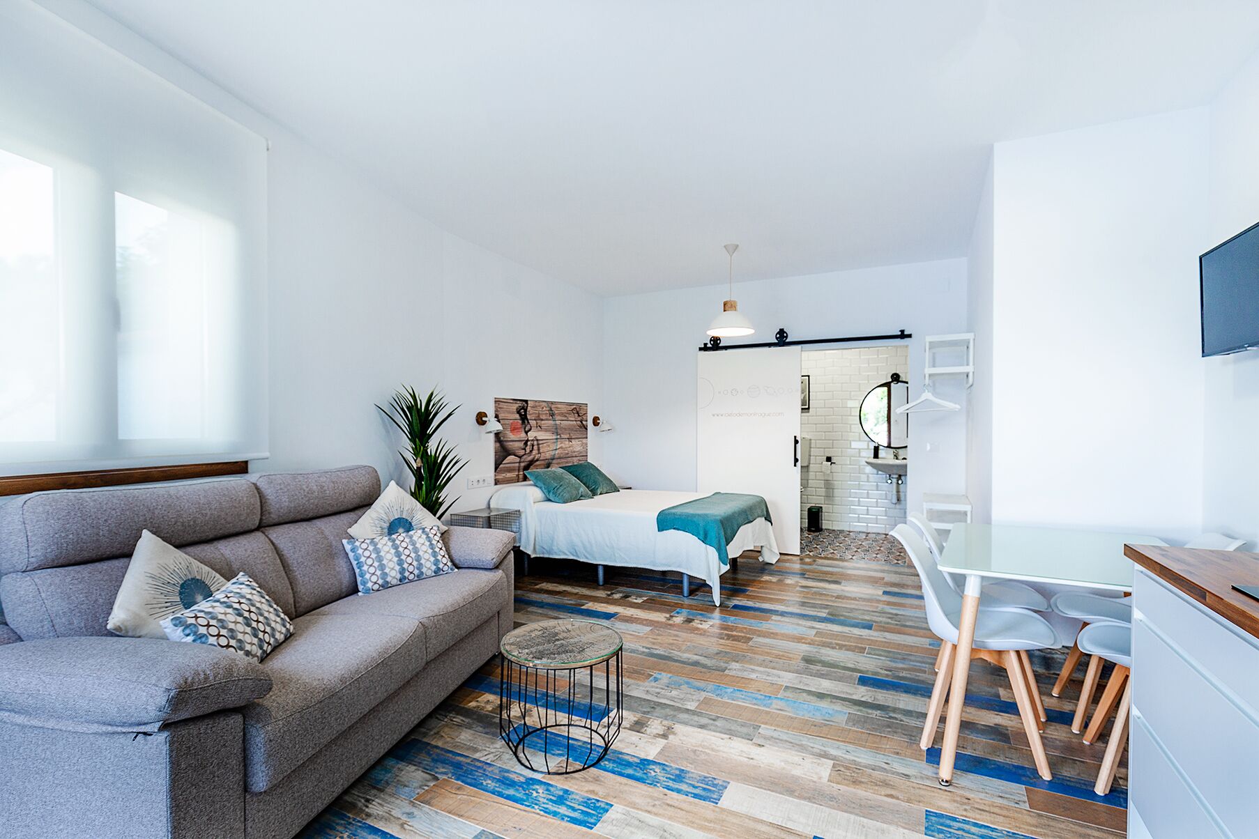 Una sala moderna con un sofá, una mesita redonda y una cama con cabecera blanca. Se ve una escalera que conduce a un baño con un lavabo y un espejo.