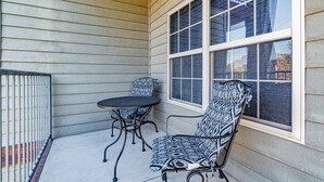 Un patio cubierto con una mesa y dos sillas estampadas, una ventana y una barandilla metálica.
