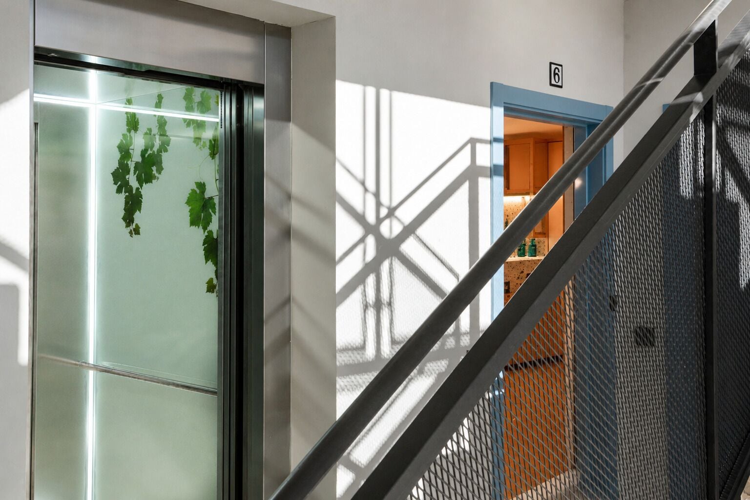 Un interno moderno con una porta in vetro, una scala in metallo e vista su una stanza con un motivo geometrico sul muro.