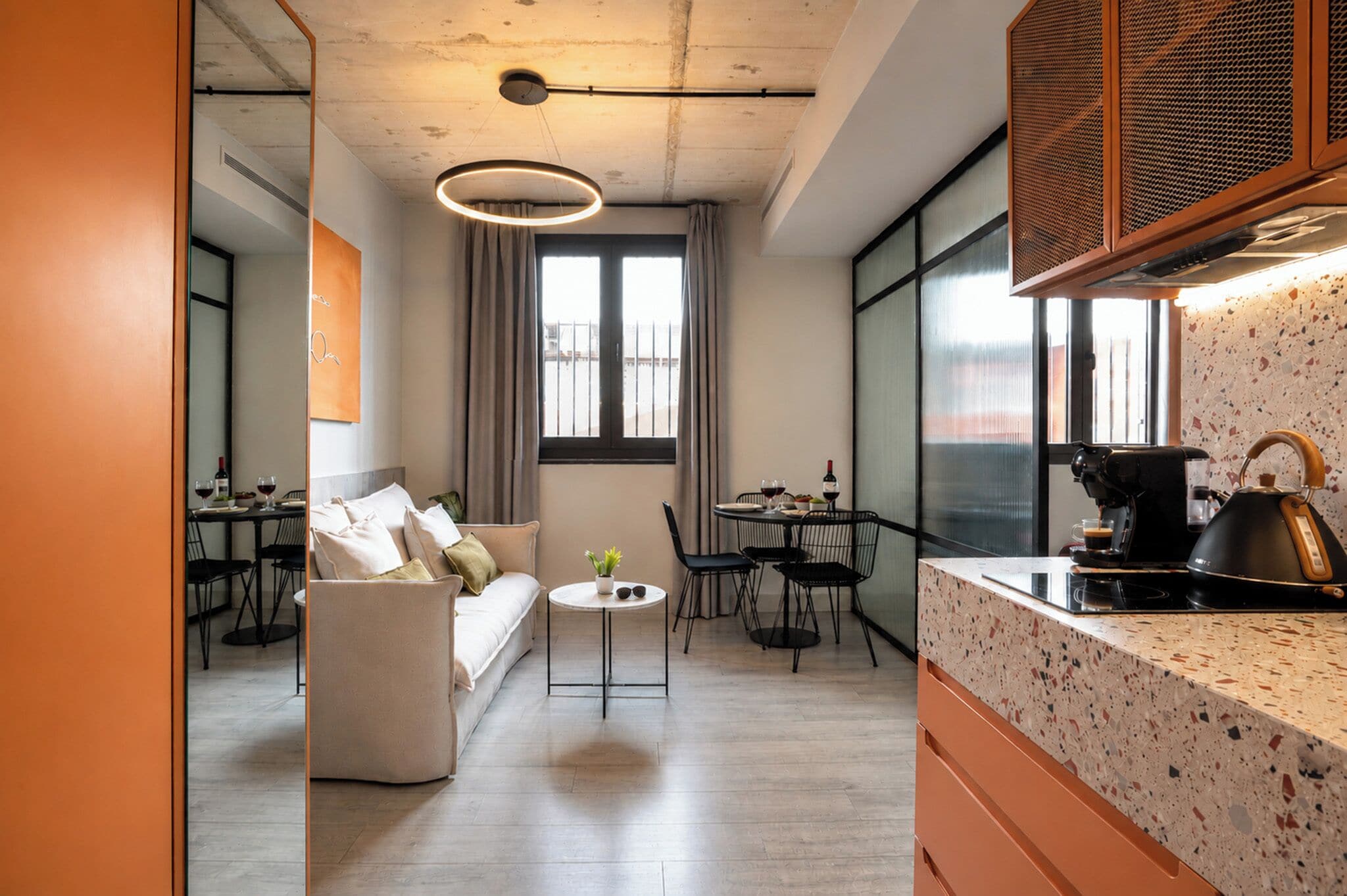 A modern living room with a sofa, a dining area with chairs, a kitchen with a kettle, and a large window with a view.