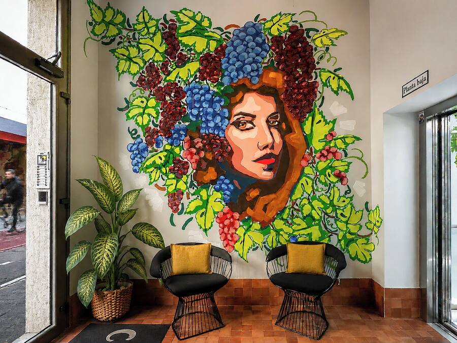 Un mural en la pared con el rostro de una mujer rodeado de parras y hojas. Hay dos sillas negras con cojines amarillos frente al mural.