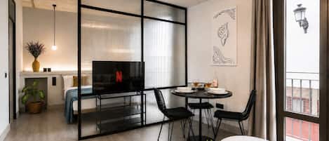 A modern living room with a TV, a dining area with chairs, a small table with a plant, and a large window with a view.