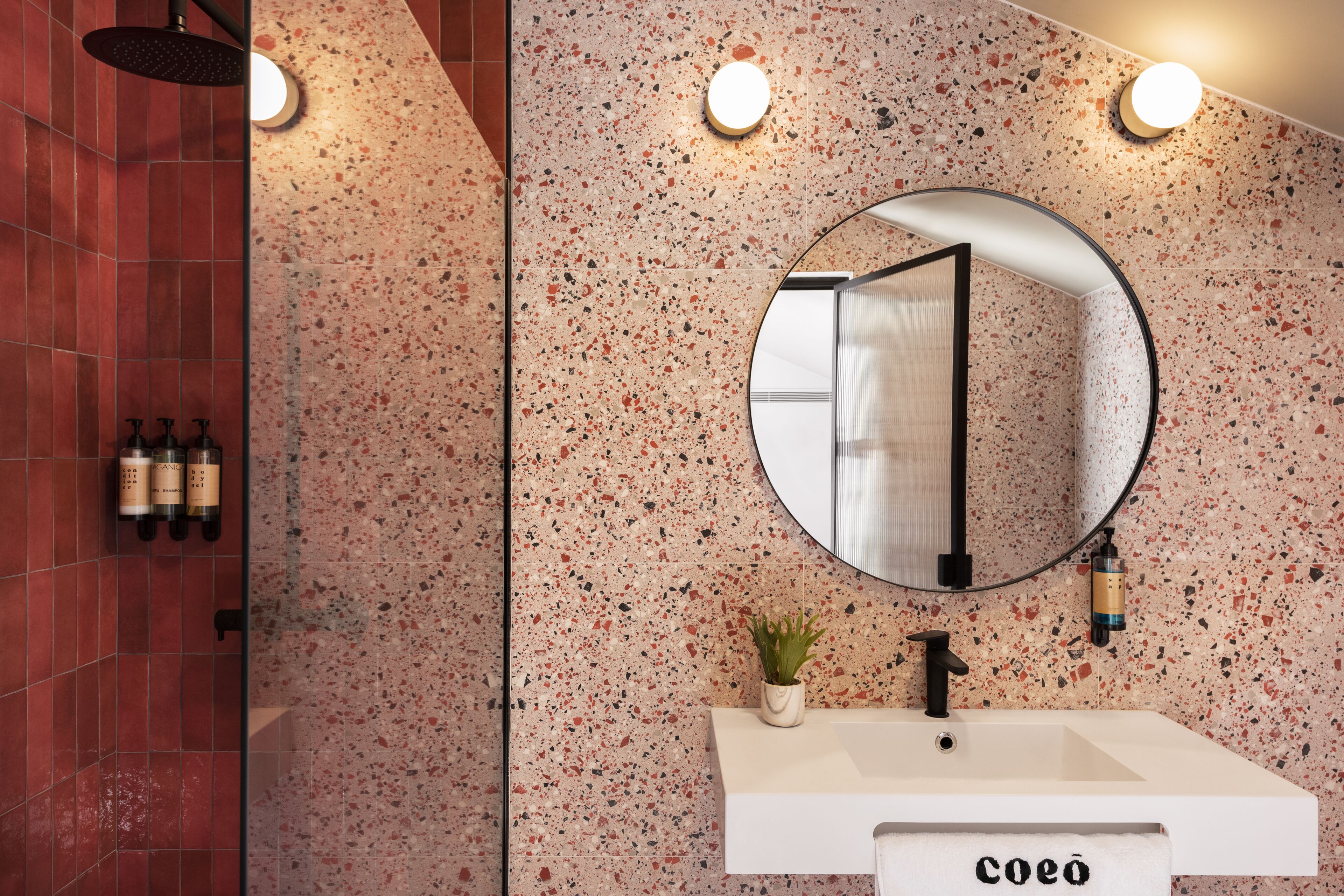 Un baño con un lavabo blanco, un espejo redondo y una ducha con pared de azulejos rojos.