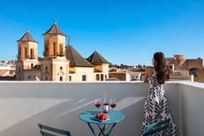 A woman on a balcony overlooking a historic building with two towers, a table with wine glasses and fruit, and a clear blue sky. - Coeo Parras Design Apartments (Málaga)