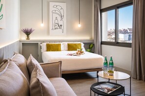 A hotel room with a bed, a sofa, a round table with drinks, and a window with a view of buildings. - Coeo Parras Design Apartments (Málaga)