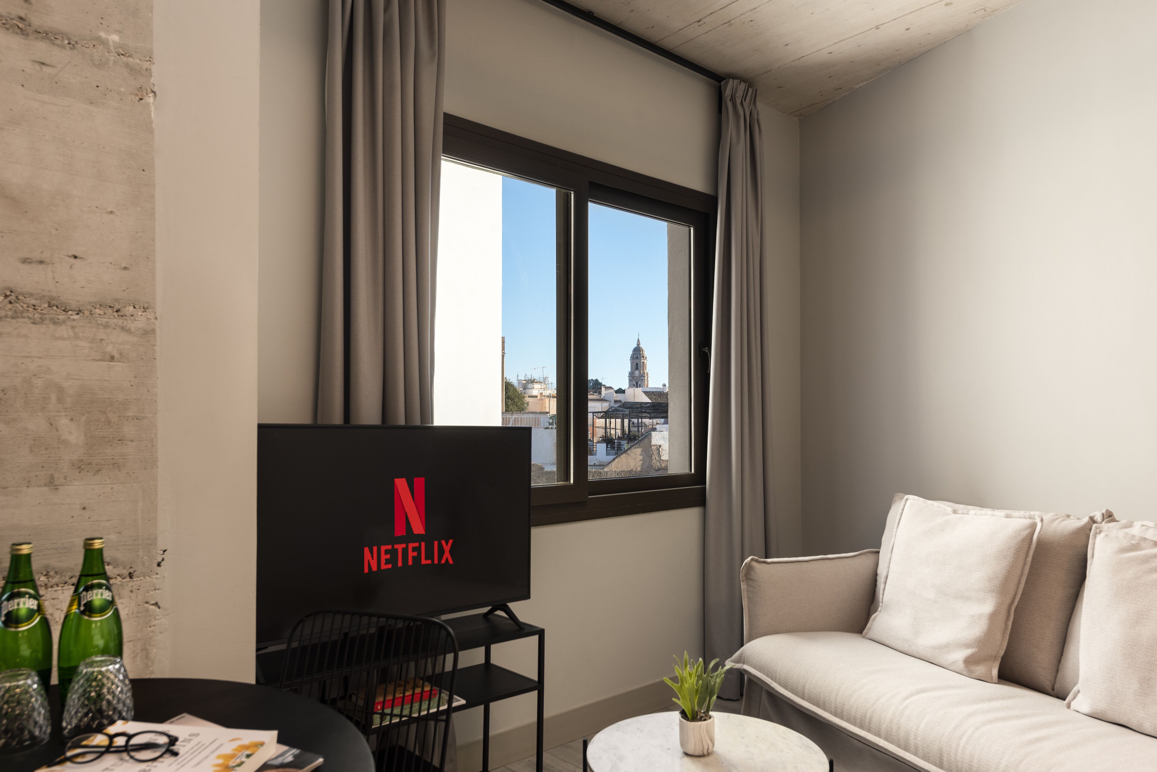 Una sala de estar moderna con un ventanal grande, un televisor de pantalla plana mostrando el logo de Netflix, un sofá, una mesa de centro y una pequeña planta.