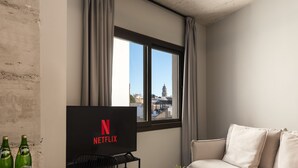 Una sala de estar moderna con un ventanal grande, un televisor de pantalla plana mostrando el logo de Netflix, un sofá, una mesa de centro y una pequeña planta.