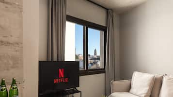 Una sala de estar moderna con un ventanal grande, un televisor de pantalla plana mostrando el logo de Netflix, un sofá, una mesa de centro y una pequeña planta.