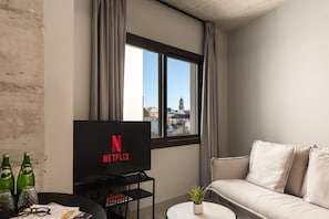A modern living room with a large window, a flat-screen TV displaying the Netflix logo, a sofa, a coffee table, and a small plant.