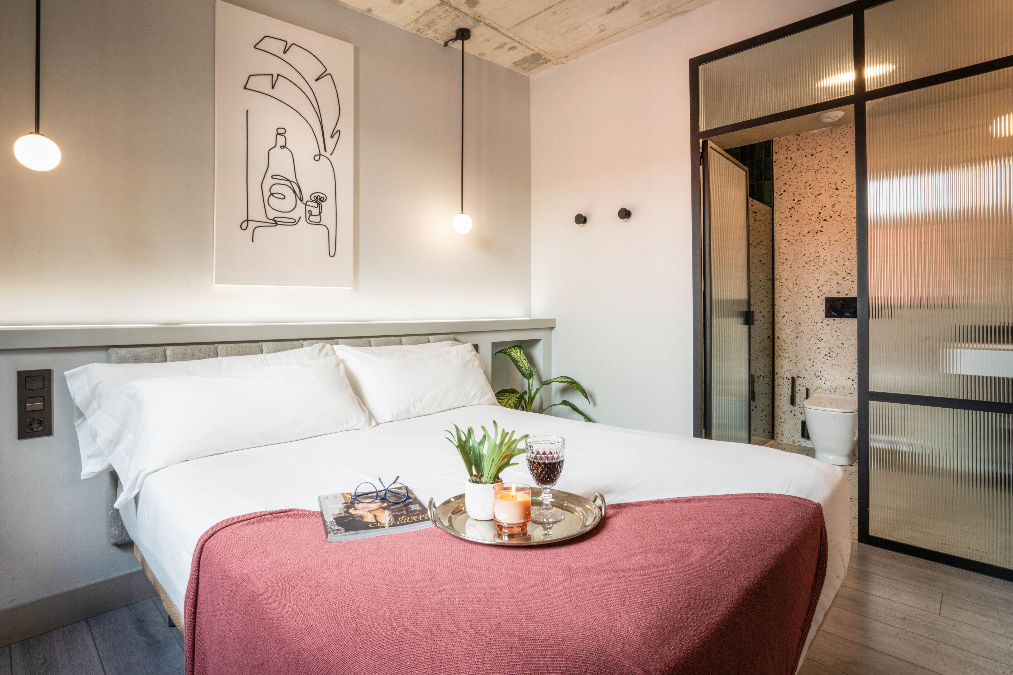 Un lit bien fait, avec un plateau contenant divers objets, une œuvre d’art accrochée au mur et une plante.