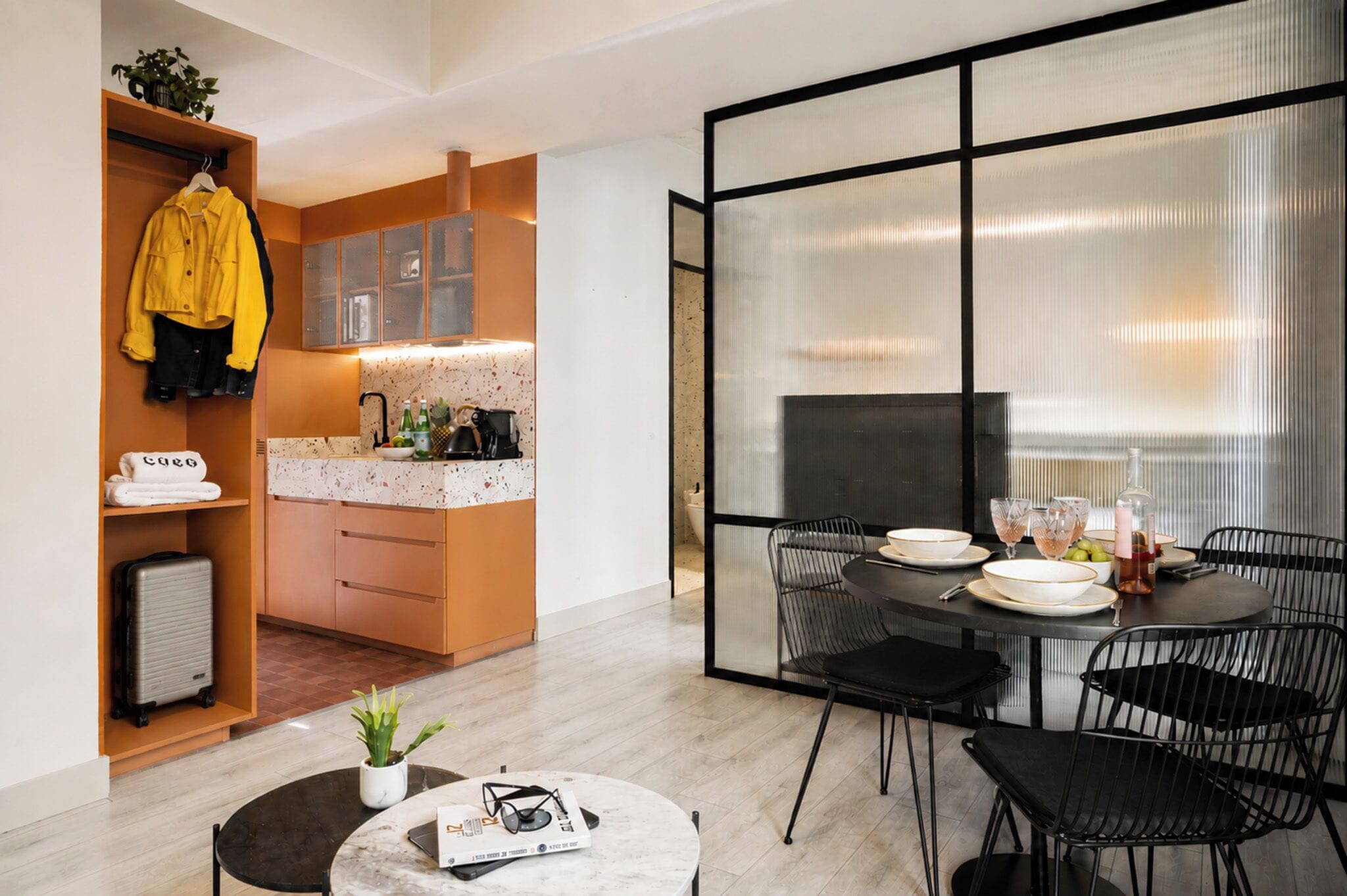 A modern kitchen with a dining area, a small table with a plant, and a yellow jacket hanging on a rack.