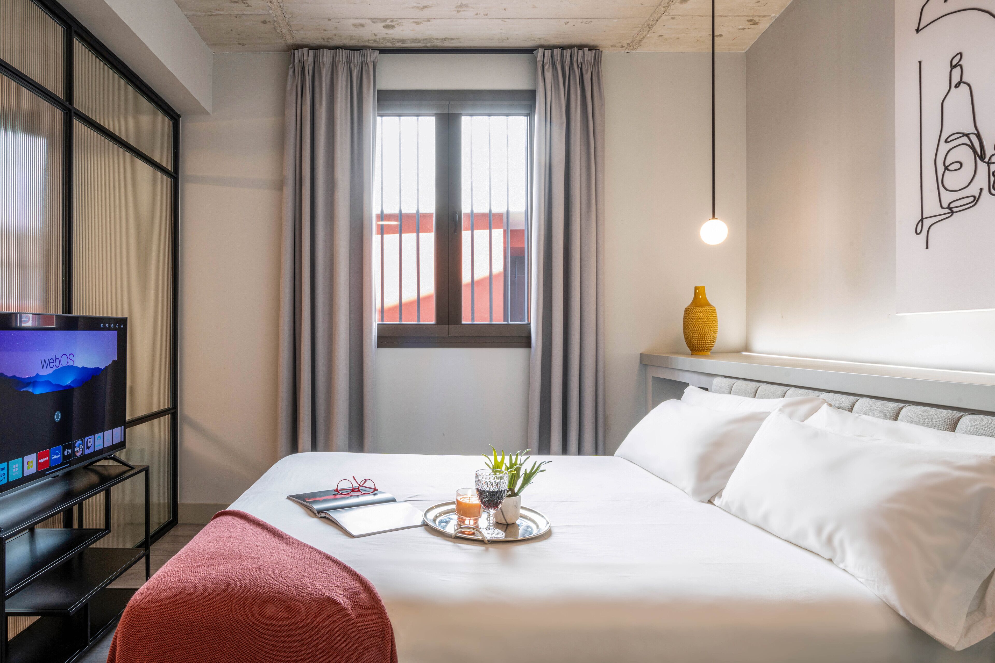 A hotel room with a bed, a TV on a stand, a window with curtains, a bedside table with a vase, and a wall art piece.