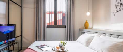 A hotel room with a bed, a TV on a stand, a window with curtains, a bedside table with a vase, and a wall art piece.