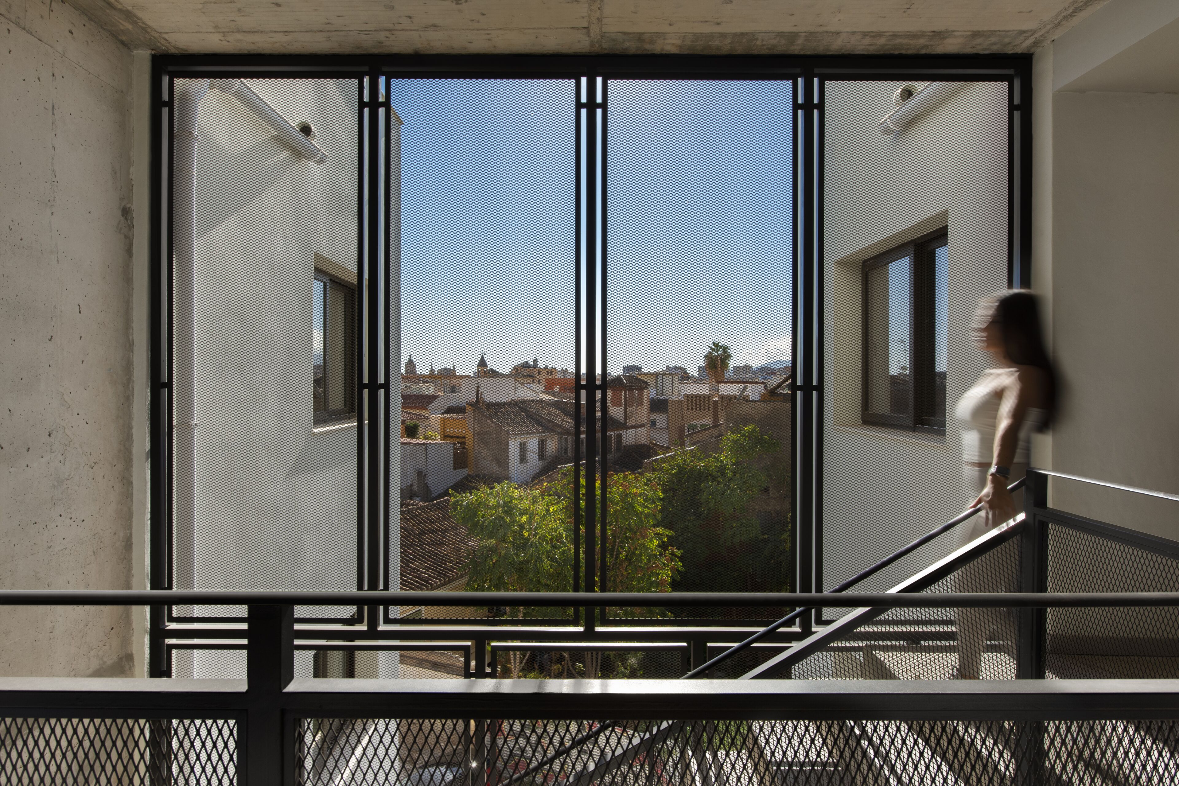 Vista a través de una ventana con reja metálica, que muestra un paisaje urbano con edificios y zonas verdes.