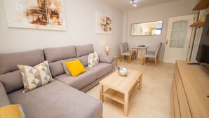 A modern living room with a grey sofa, a wooden coffee table, and a dining area with chairs and a table. - Mikonos Playa apto 15 (Manilva)