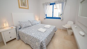 A neatly arranged bedroom with a bed, bedside tables, a chair, and a window with curtains. - Mikonos Playa apto 15 (Manilva)