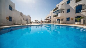 A swimming pool with clear blue water, surrounded by white buildings with balconies and windows. - Mikonos Playa apto 15 (Manilva)