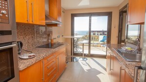Une cuisine compacte dotée de comptoirs en granit, d’appareils électroménagers en acier inoxydable et d’armoires en bois. Une porte-fenêtre coulissante donne accès à un balcon aménagé avec des sièges d’extérieur.