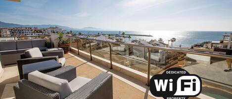 A rooftop terrace with wicker furniture and a glass railing overlooking a coastal area.